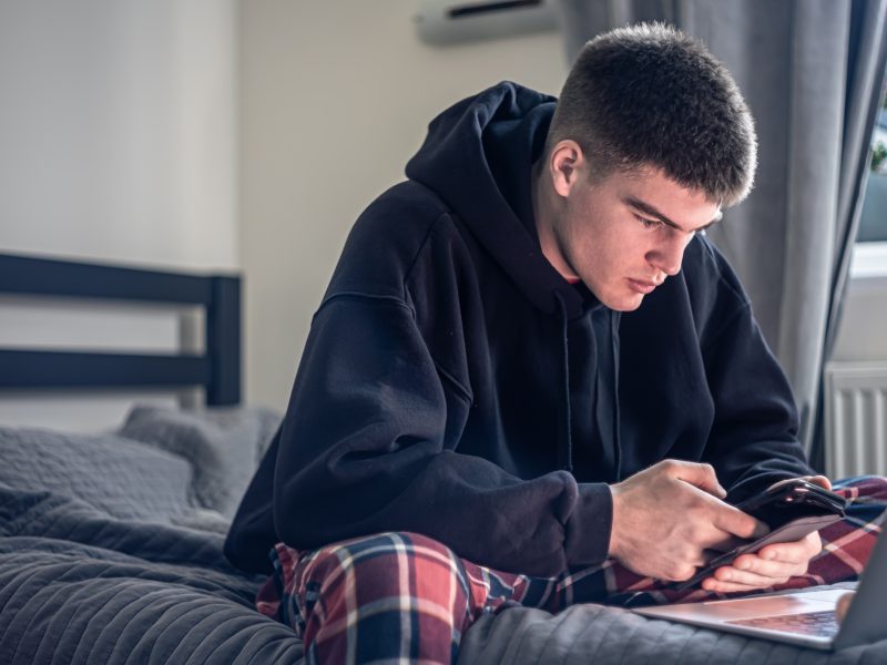 A teenager guy sits in a room on a bed and uses a laptop.