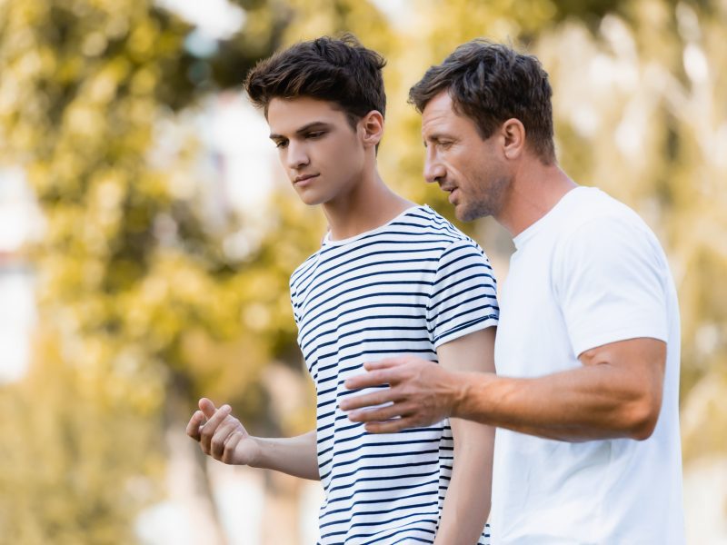 father gesturing and talking with teenager son in park